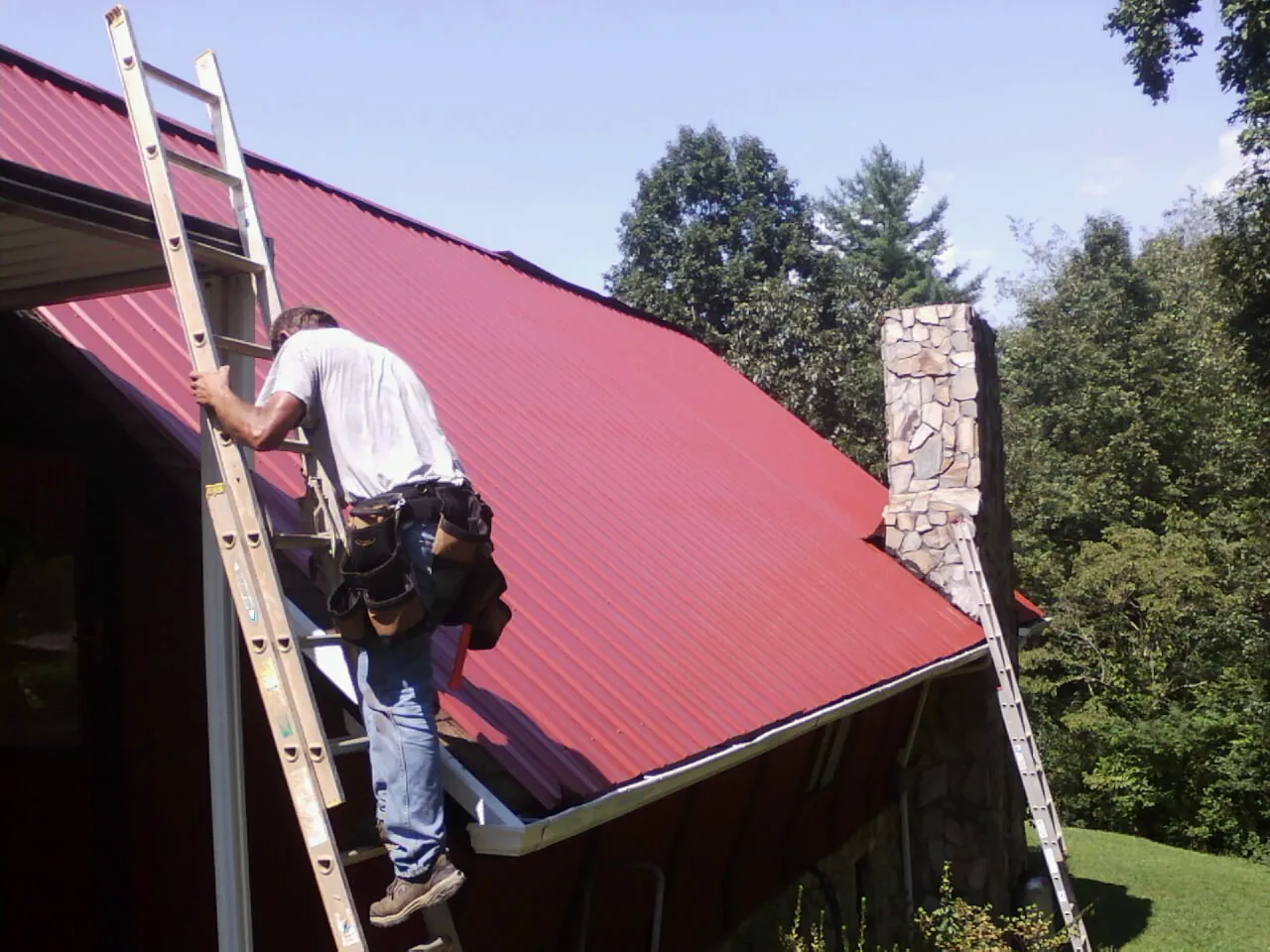 Roofer on a ladder beside a red metal roof and stone chimney — Soffit & Fascia Repair by Roofing Brothers in Shavertown
